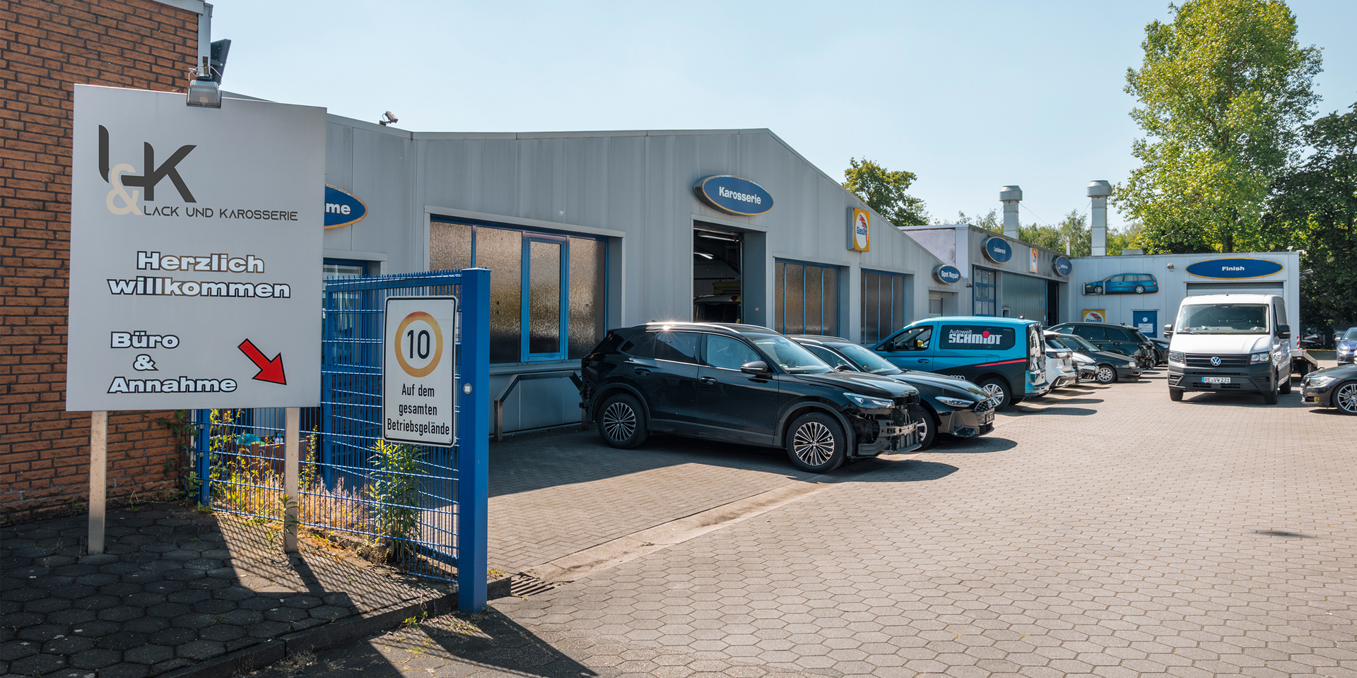 „Außenansicht des Standorts Lack und Karosserie von Autowelt Schmidt mit Werkstattgebäude, Einfahrt, geparkten Fahrzeugen und Hinweisschild zum Büro und zur Annahme